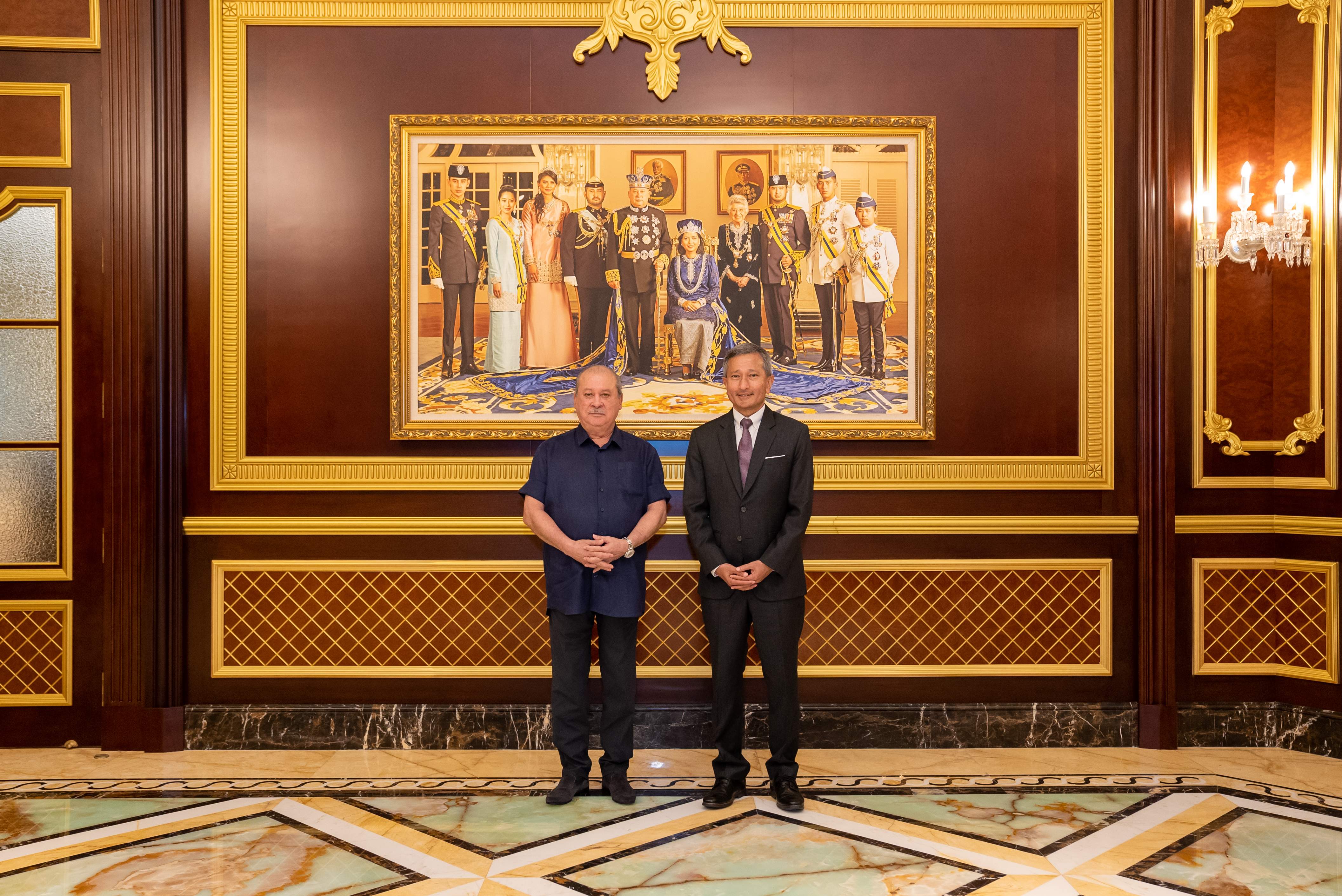 Two men stand before a large framed painting of the Brunei royal family.